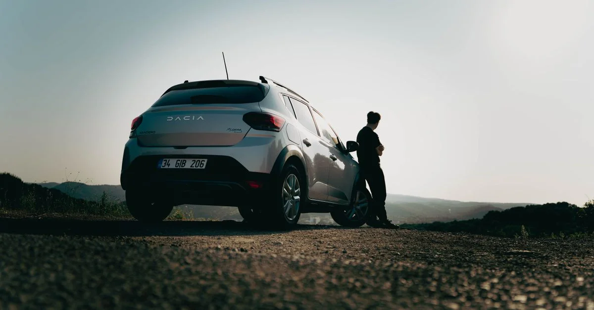 Un homme s'appuie contre une voiture Dacia sur une route pittoresque au coucher du soleil, capturant un sentiment d'envie de voyager.